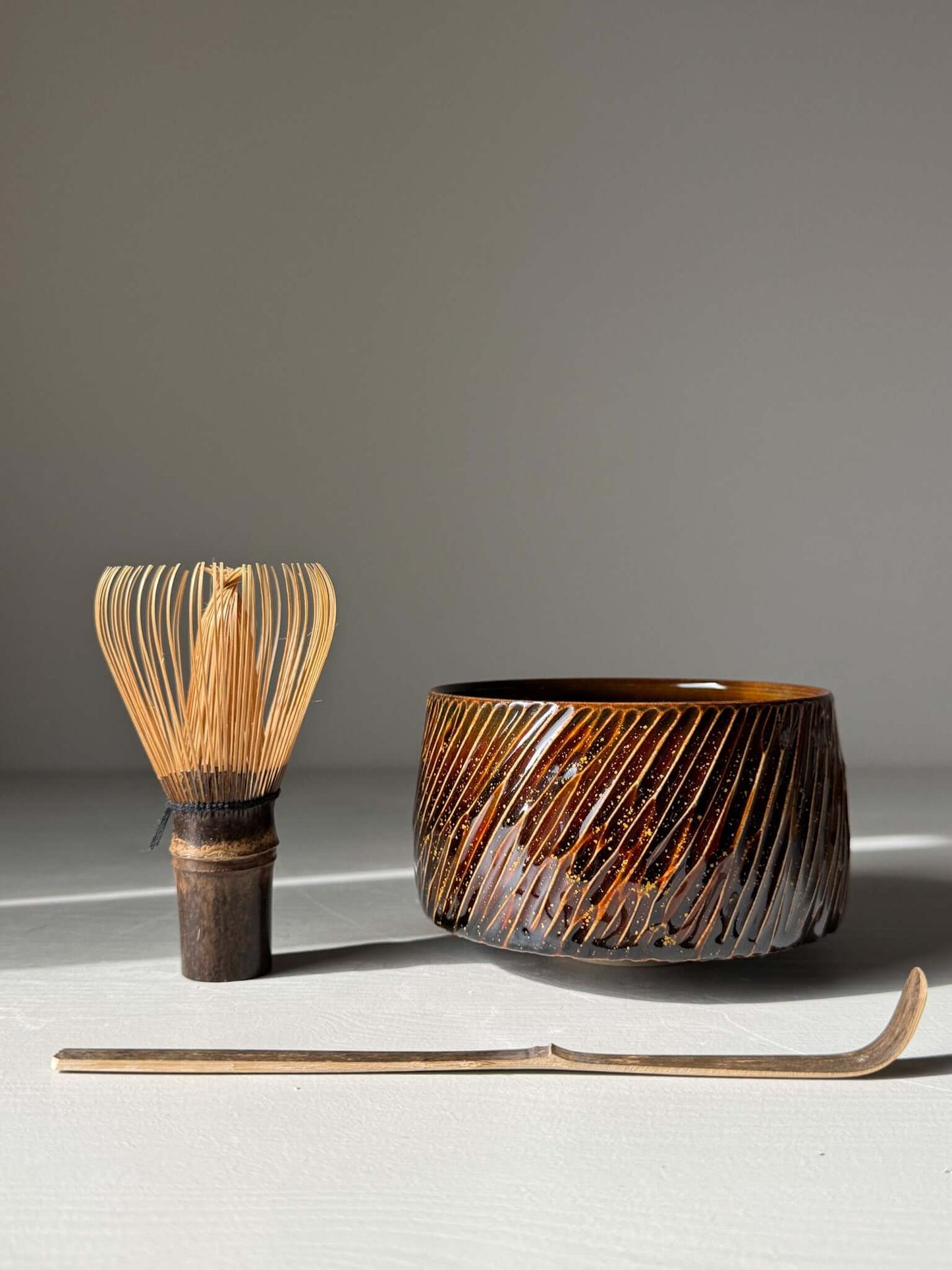 Handmade ceramic Matcha Bowl – Kinsei #04 with glossy brown glaze, shown with a bamboo whisk and scoop, evoking a galaxy-like glow.