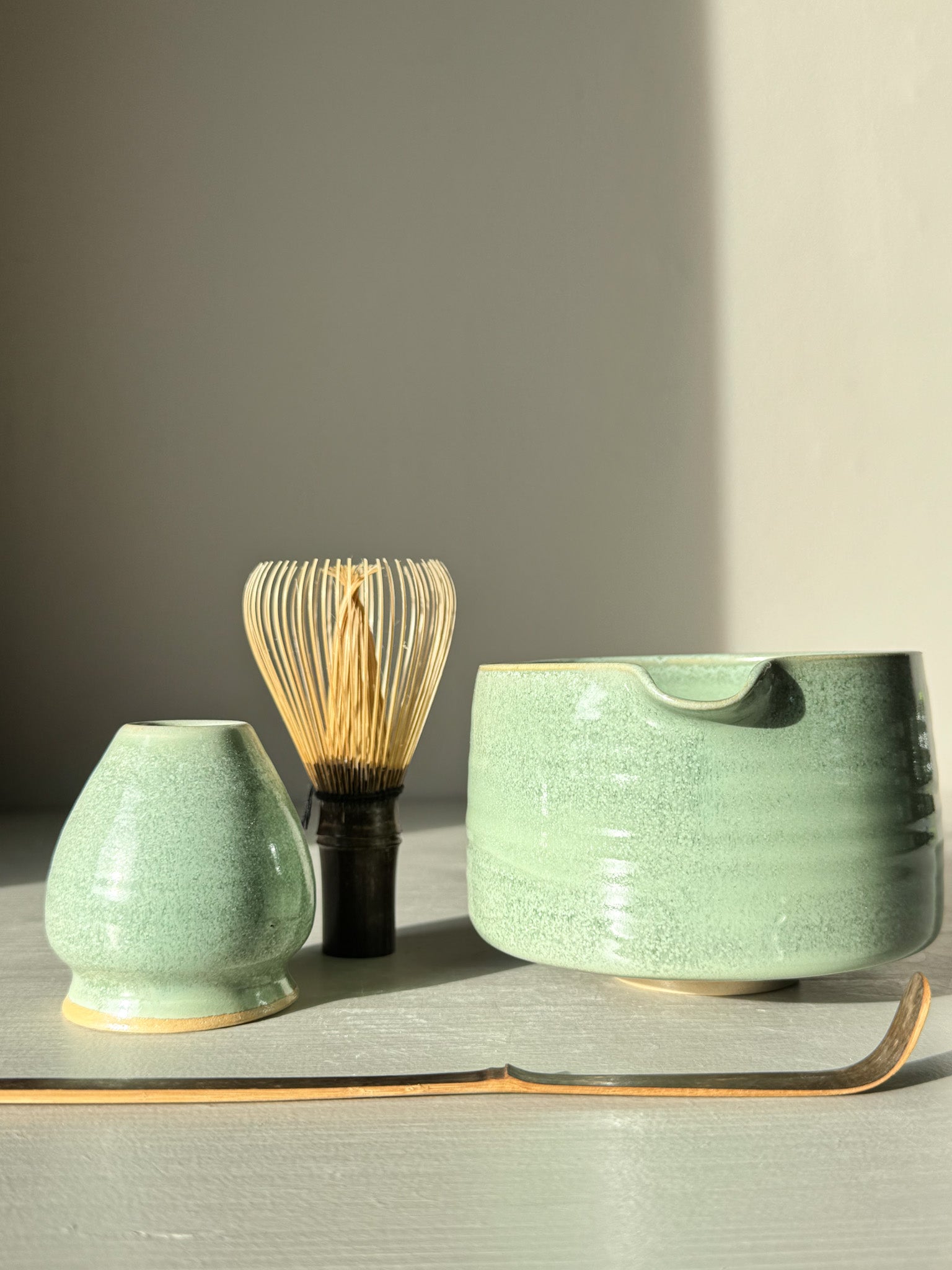 Matcha Bowl Spout – Sage #02 with bamboo whisk and spout resting on a table, showcasing its soft sage-green glaze in sunlight.