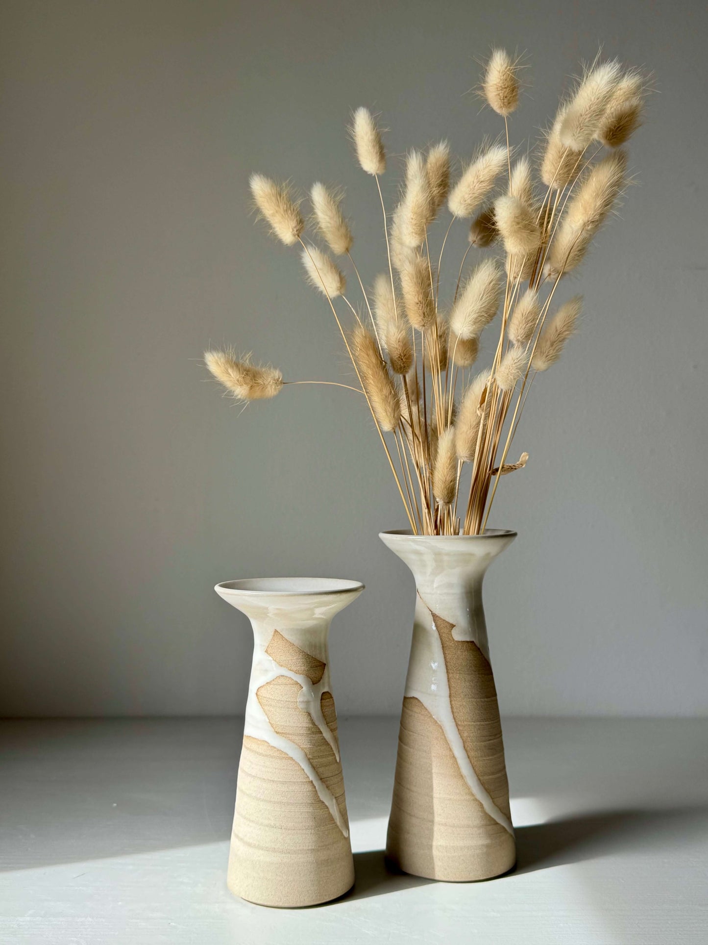 Pair of tall handmade ceramic vases with dried grasses on a white surface.