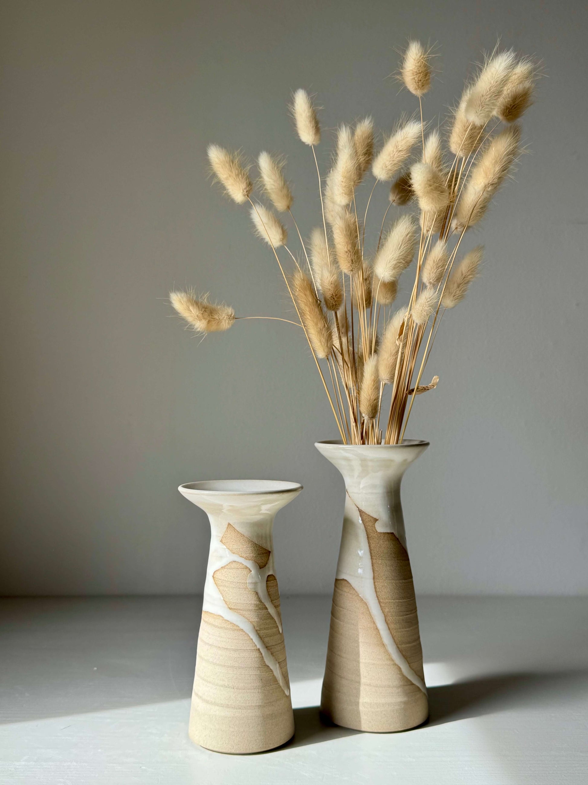 Pair of tall handmade ceramic vases with dried grasses on a white surface.