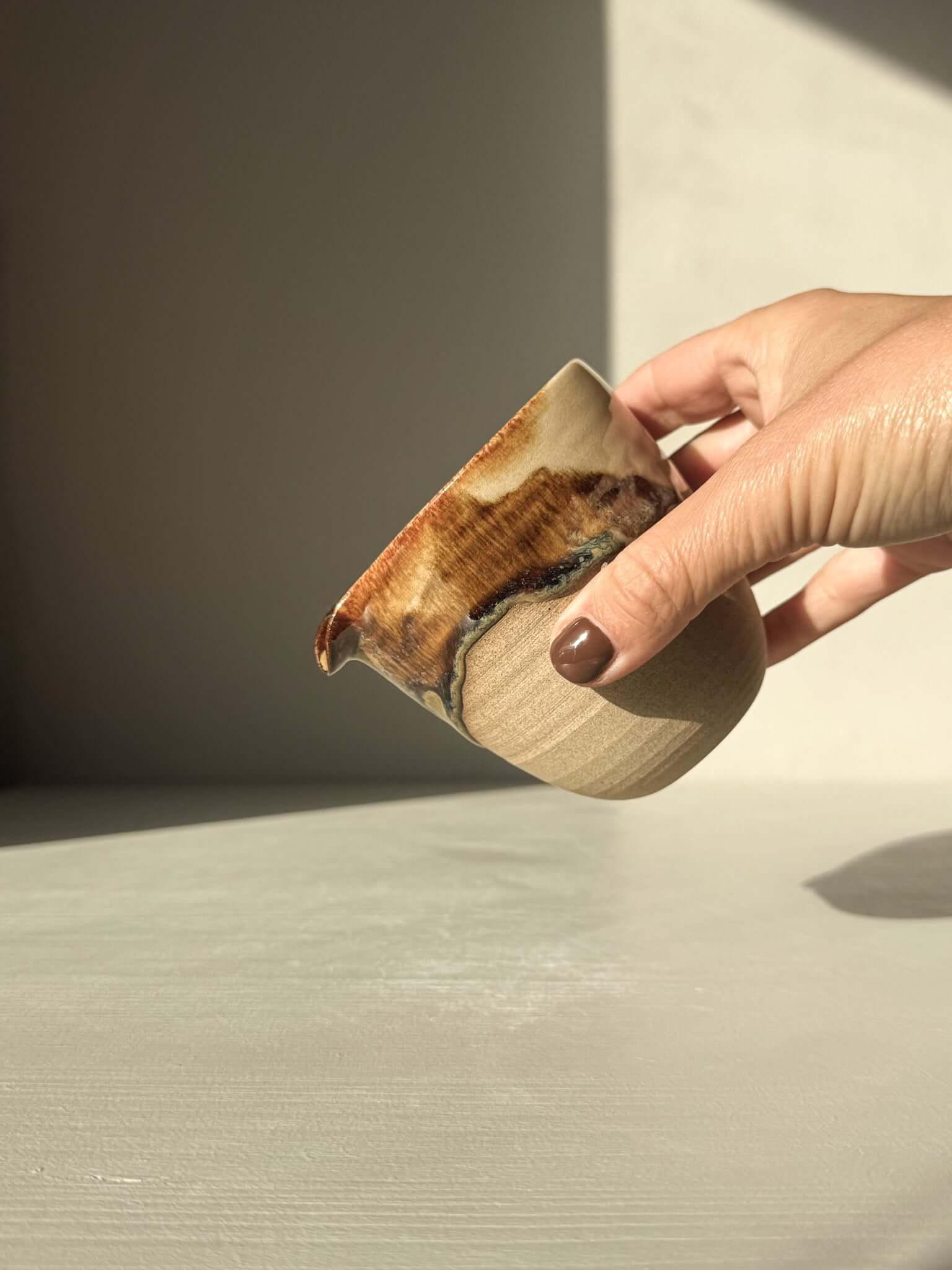 Hand holding handmade ceramic Pouring Jug – Inyo Series #01 with glossy tenmoku and clear glaze over toffee clay background