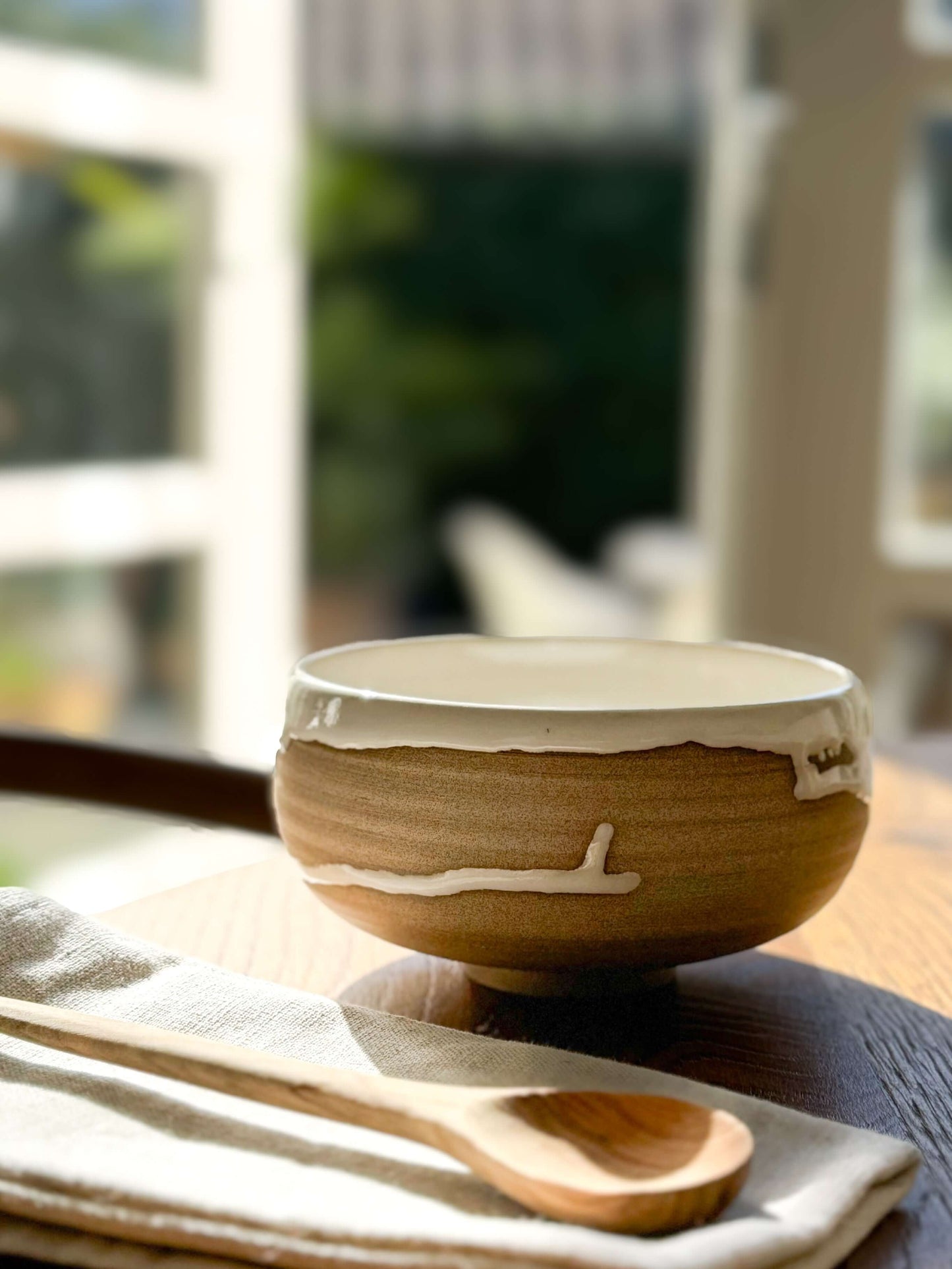 Handmade ceramic Rounded Breakfast Bowl – Yuki with toffee clay and glossy white glaze on a wooden table with a spoon.
