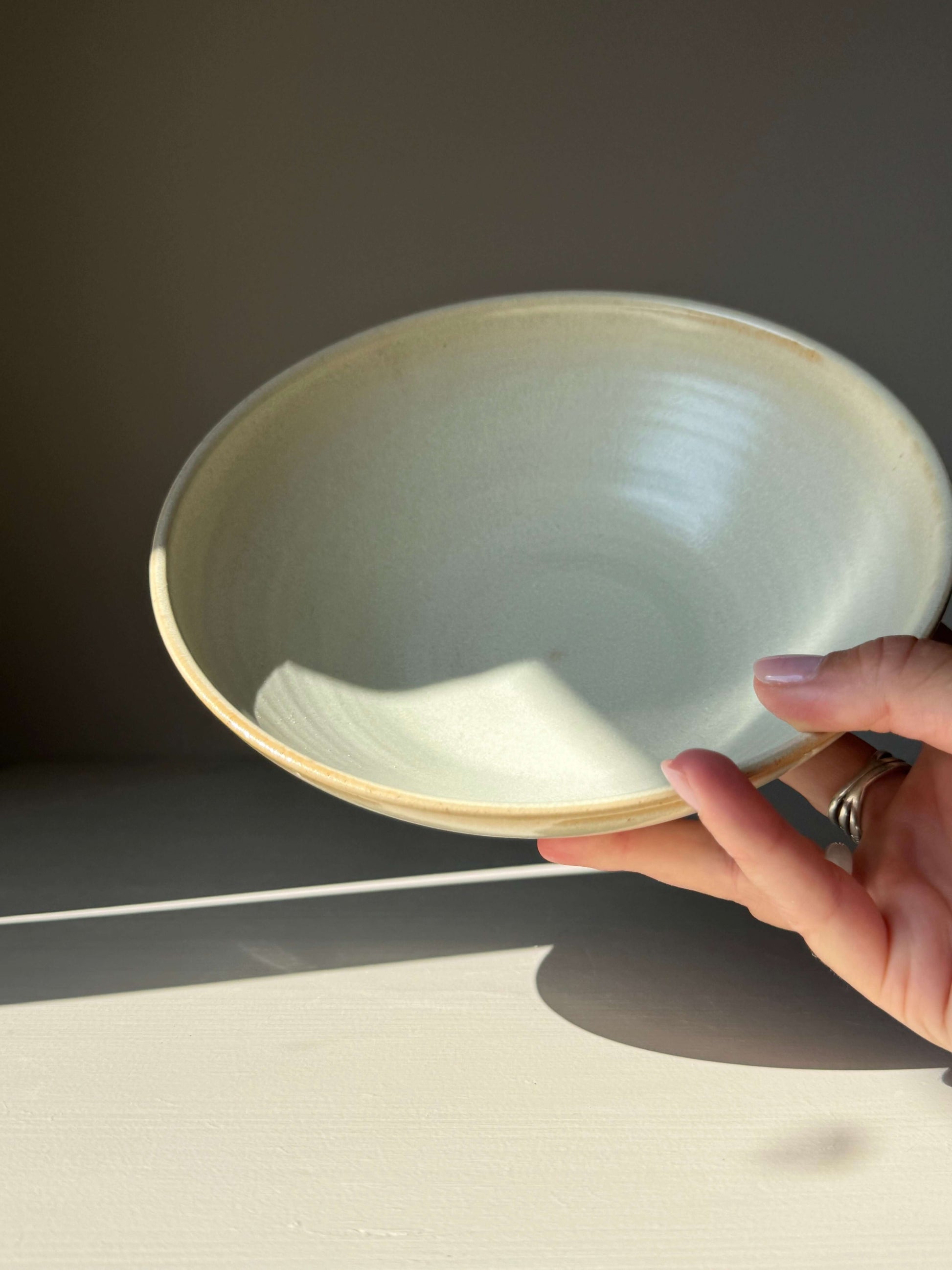Hand holding a handmade ceramic Shallow Dish - Moonstone with moonstone glaze, showcasing its elegant design on a shaded surface.