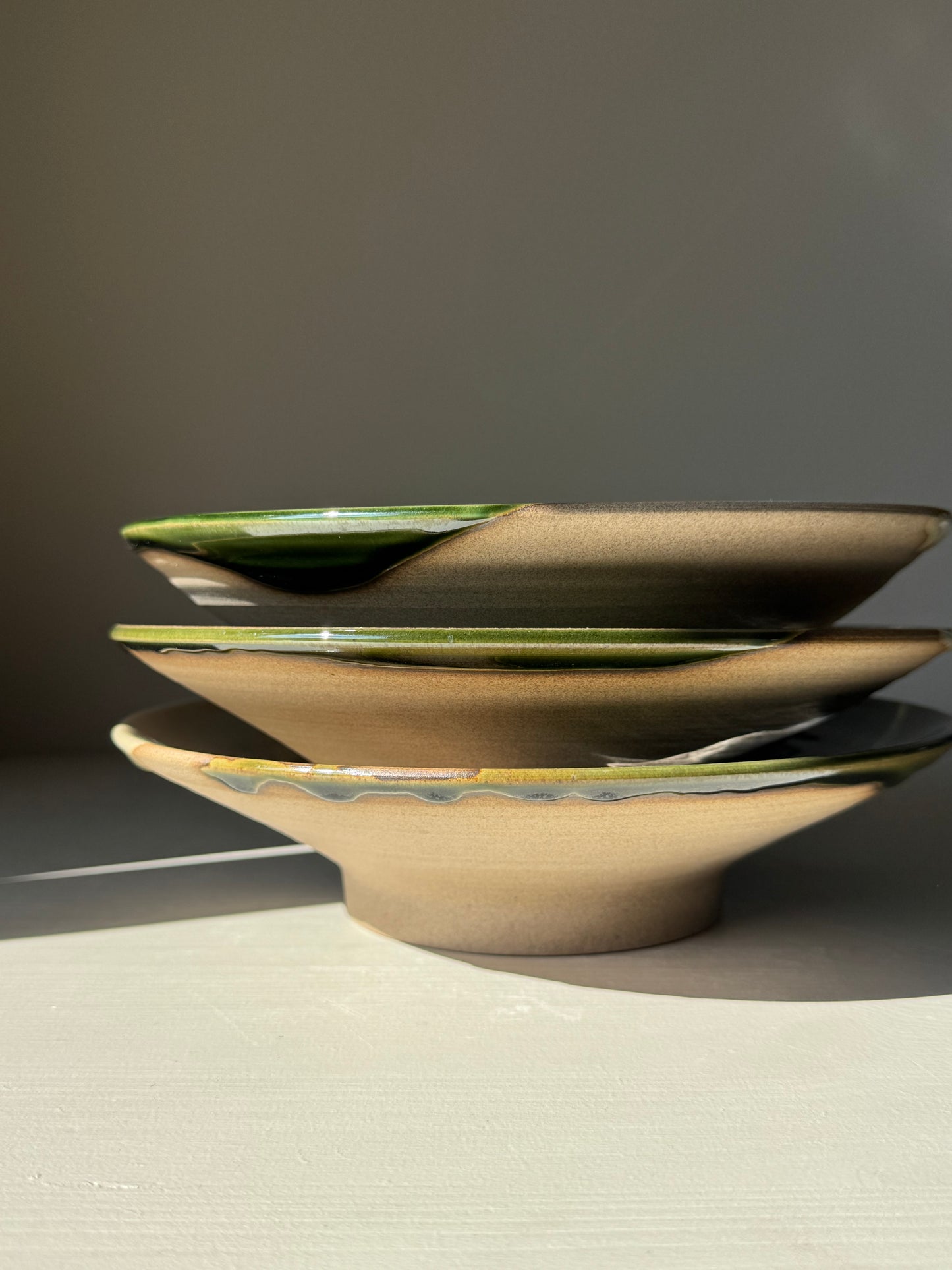 Stack of handmade ceramic Shallow Plate – Mori with forest-green finish and natural clay highlights in soft light.