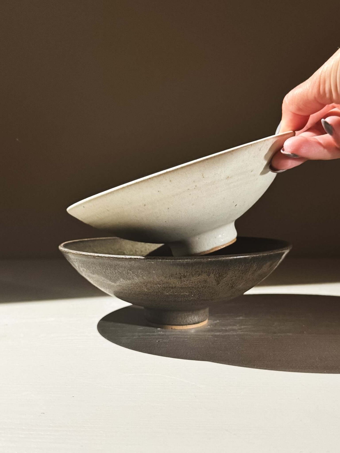 Small Footed Serving Bowl – Eggshell being held above a dark bowl, showcasing its elegant shape and matte finish.