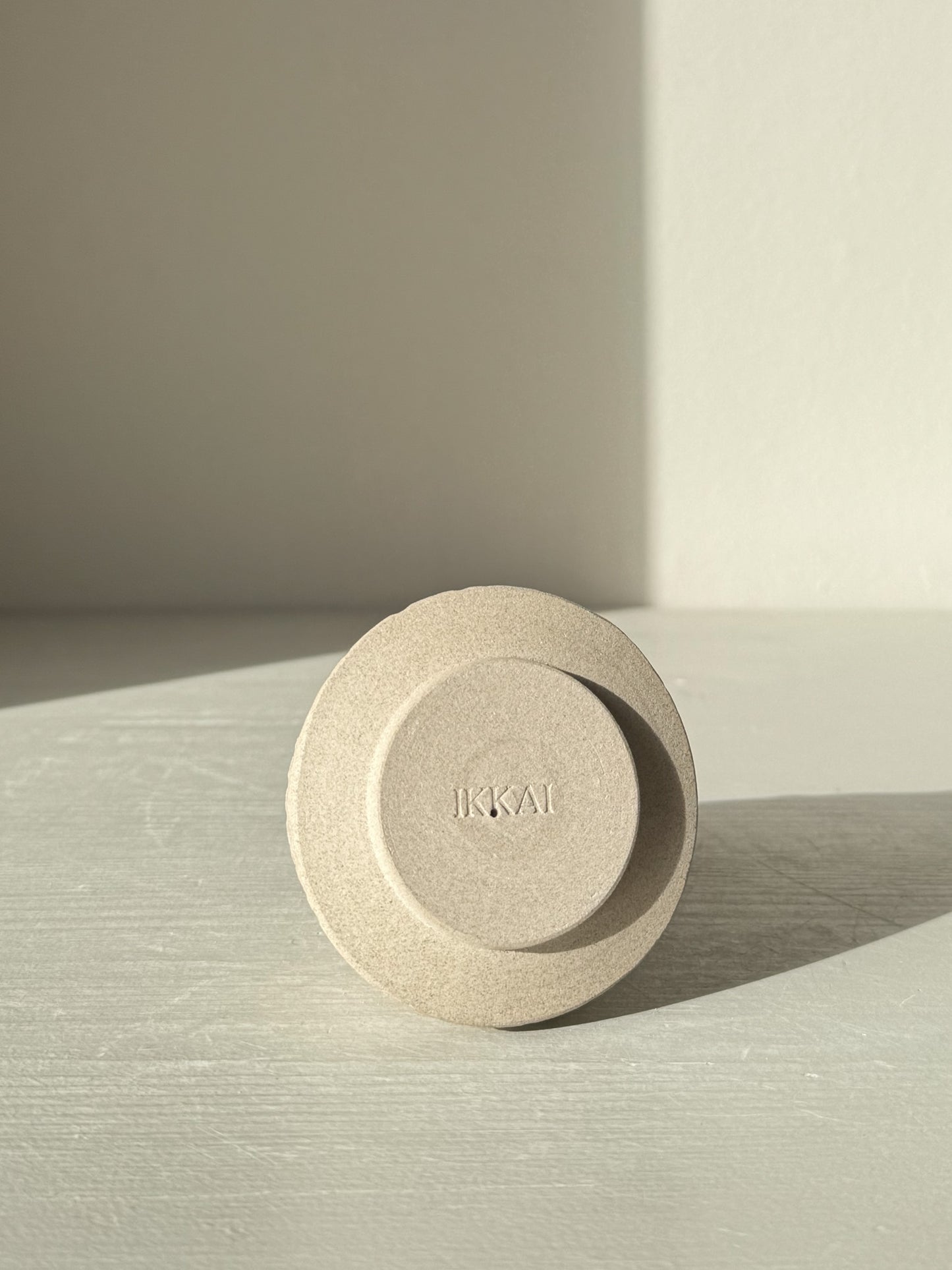 Minimalist ceramic object with engraved letters 'IKKAI', casting shadow on light surface