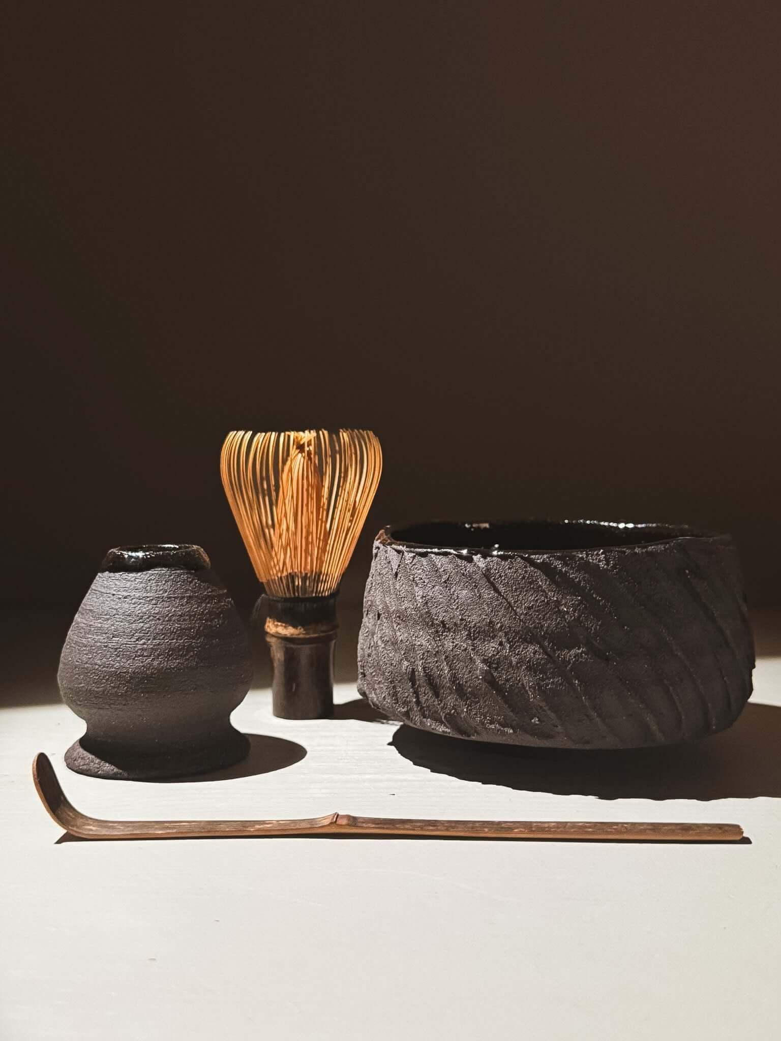 Kuro Matcha Bowl #03 with textured dark brown exterior, glossy interior, and tea whisk set against an earthy background.