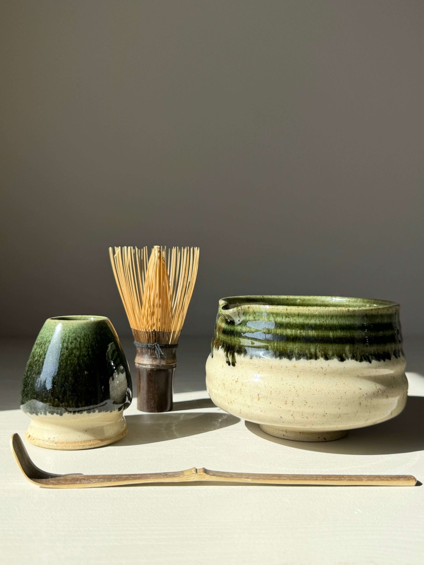 Matcha bowl with spout and green rim, accompanied by a bamboo whisk and green-glazed pottery, set against a neutral background.