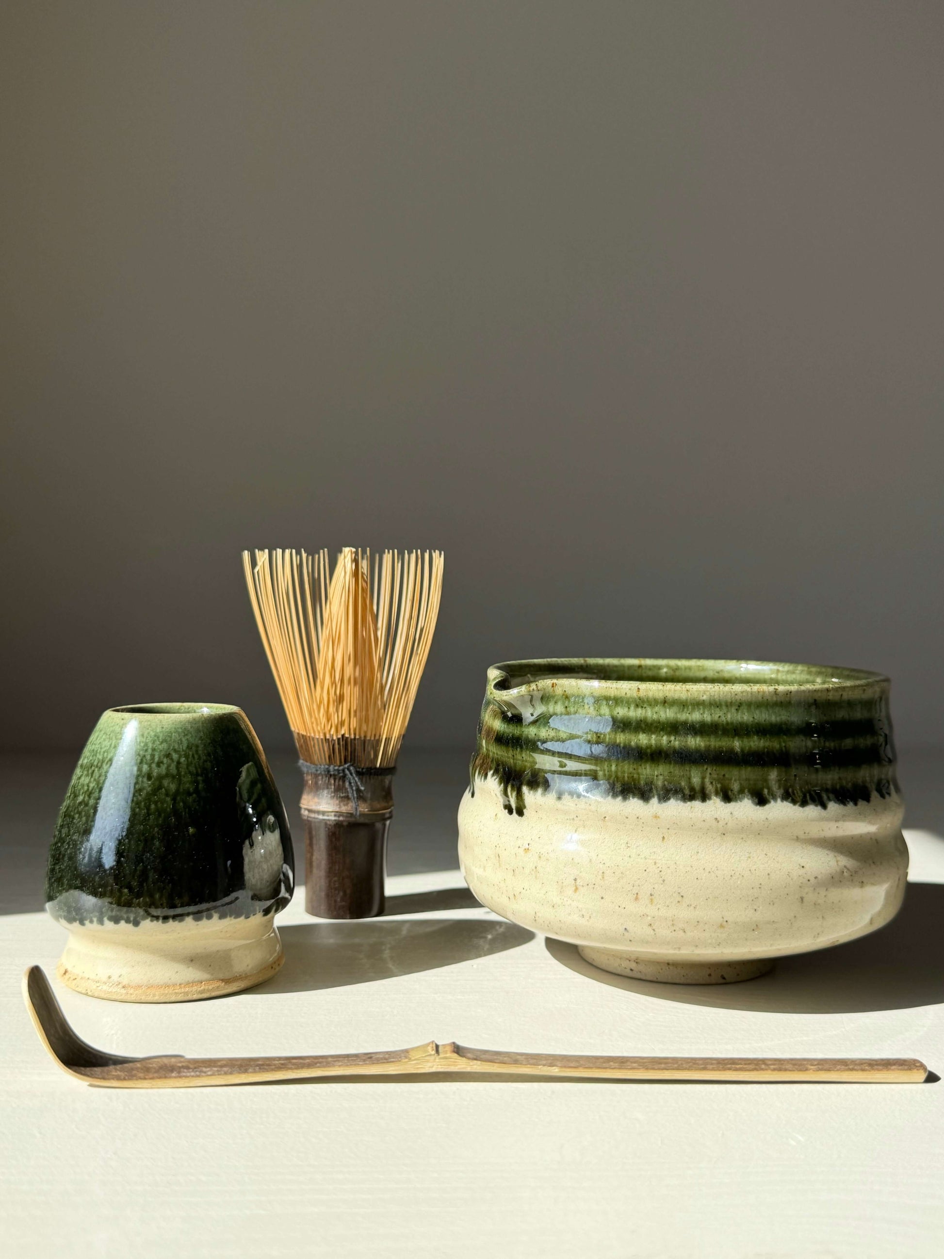 Matcha bowl with spout and green rim, accompanied by a bamboo whisk and green-glazed pottery, set against a neutral background.