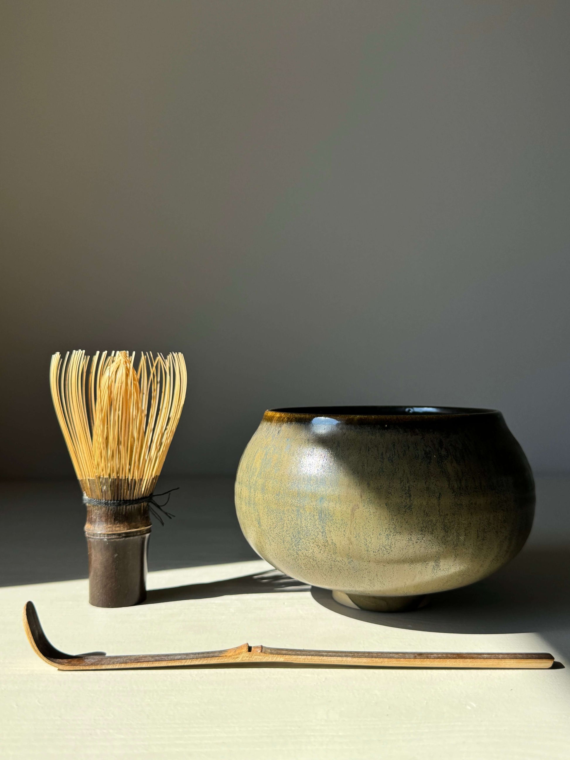 Sabi Black Matcha Bowl with bamboo whisk and scoop in soft light, featuring golden ochre and subtle blue-green tones.