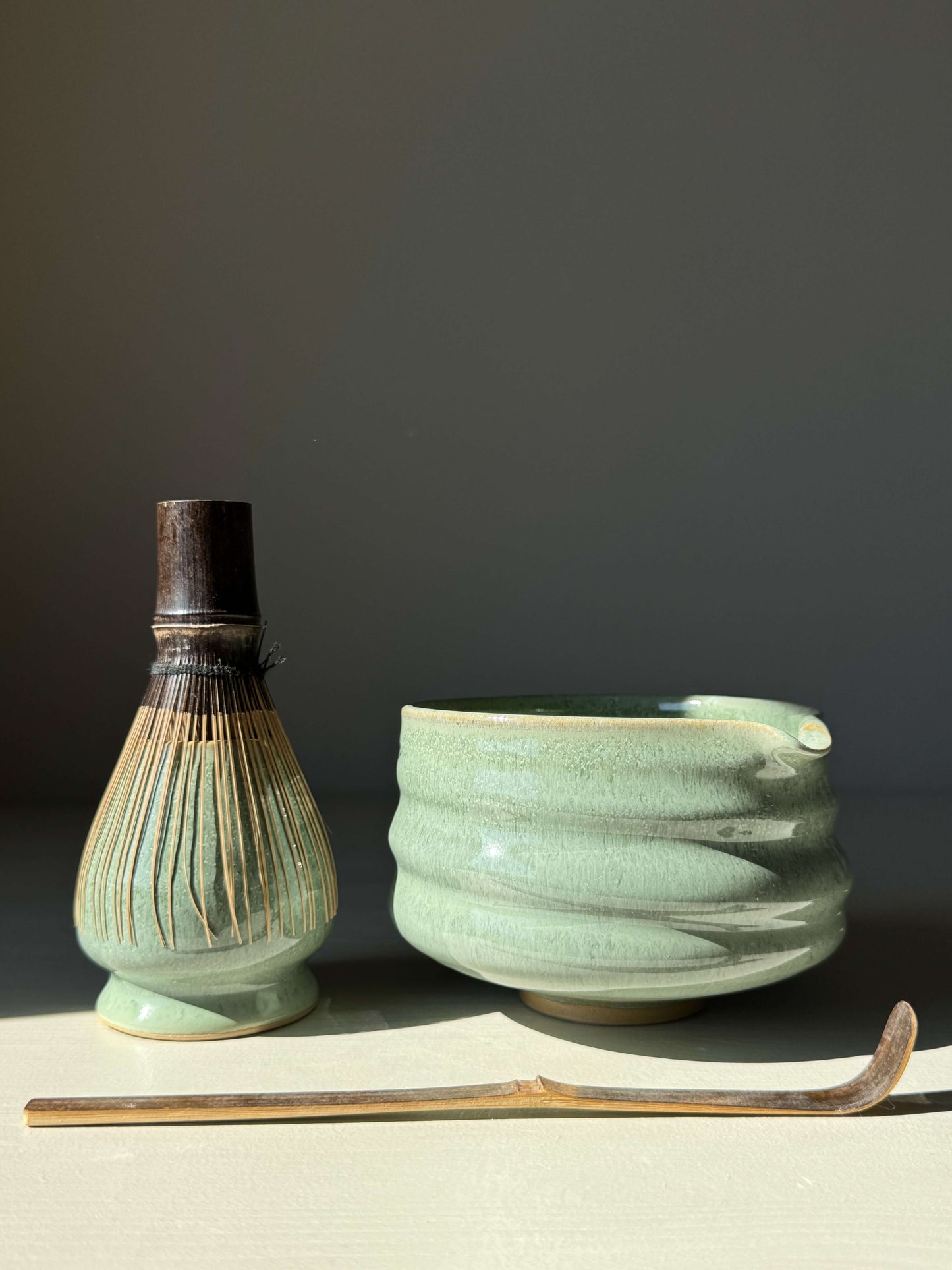 Matcha bowl with spout in sage glaze, paired with a bamboo whisk and scoop, sunlight highlighting glossy finish and ridged design.