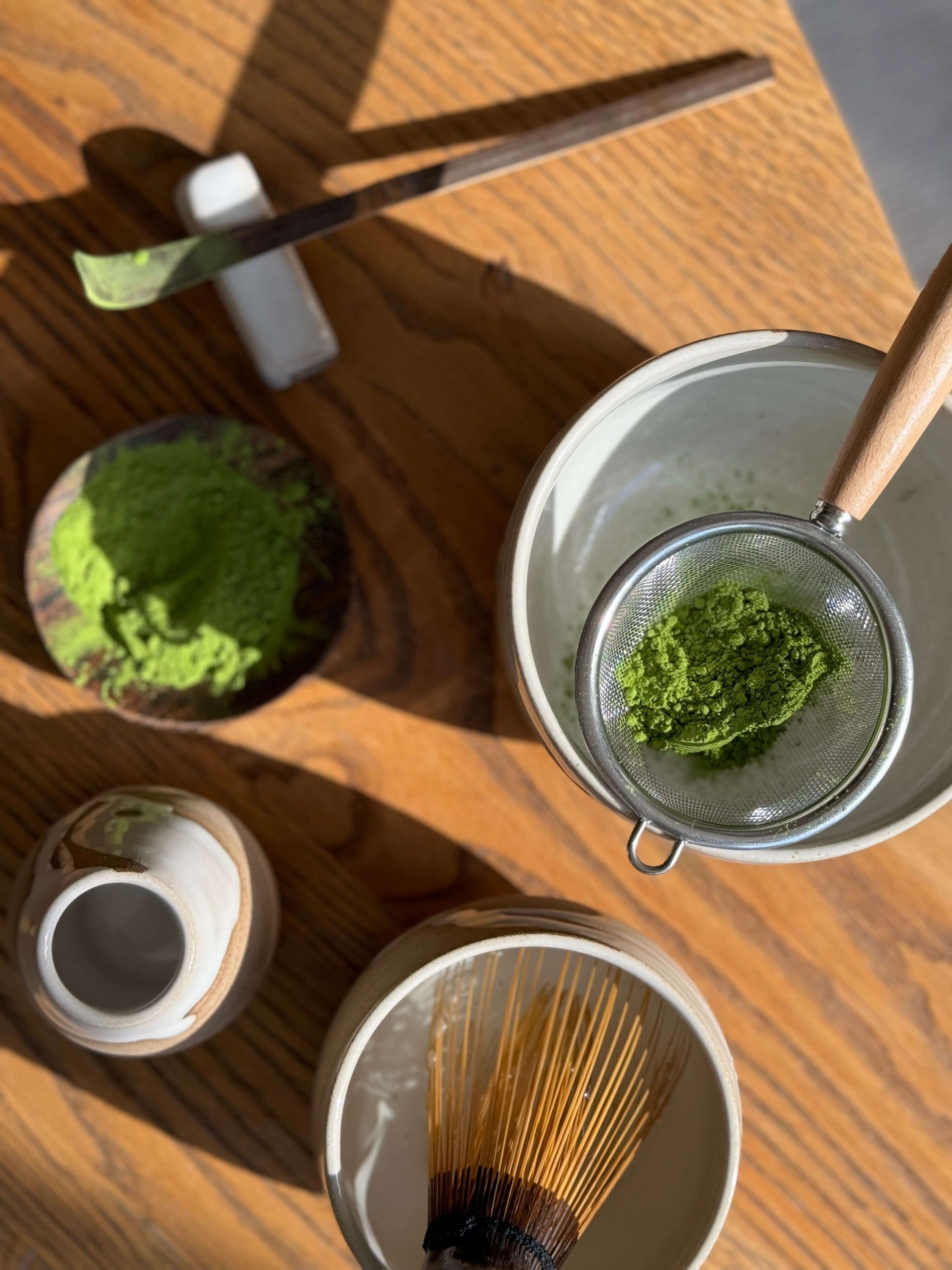 Top view of a Matcha tea set with a Yuki Series Matcha bowl, bamboo whisk, green matcha powder and sifter on a wooden table.