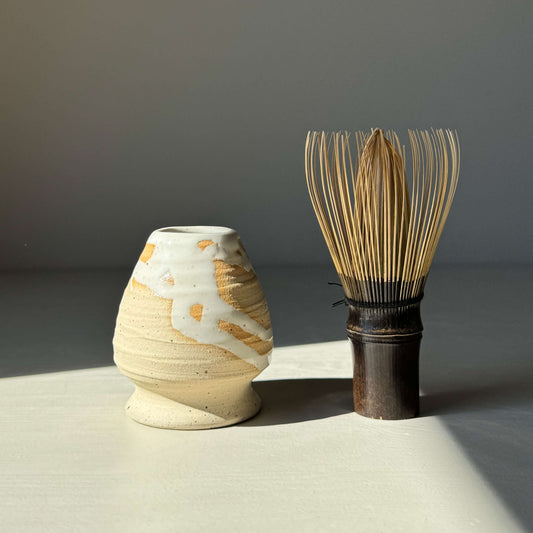 Speckled stoneware matcha whisk holder with half glossy white glaze next to bamboo matcha whisk, showcasing elegant traditional design.
