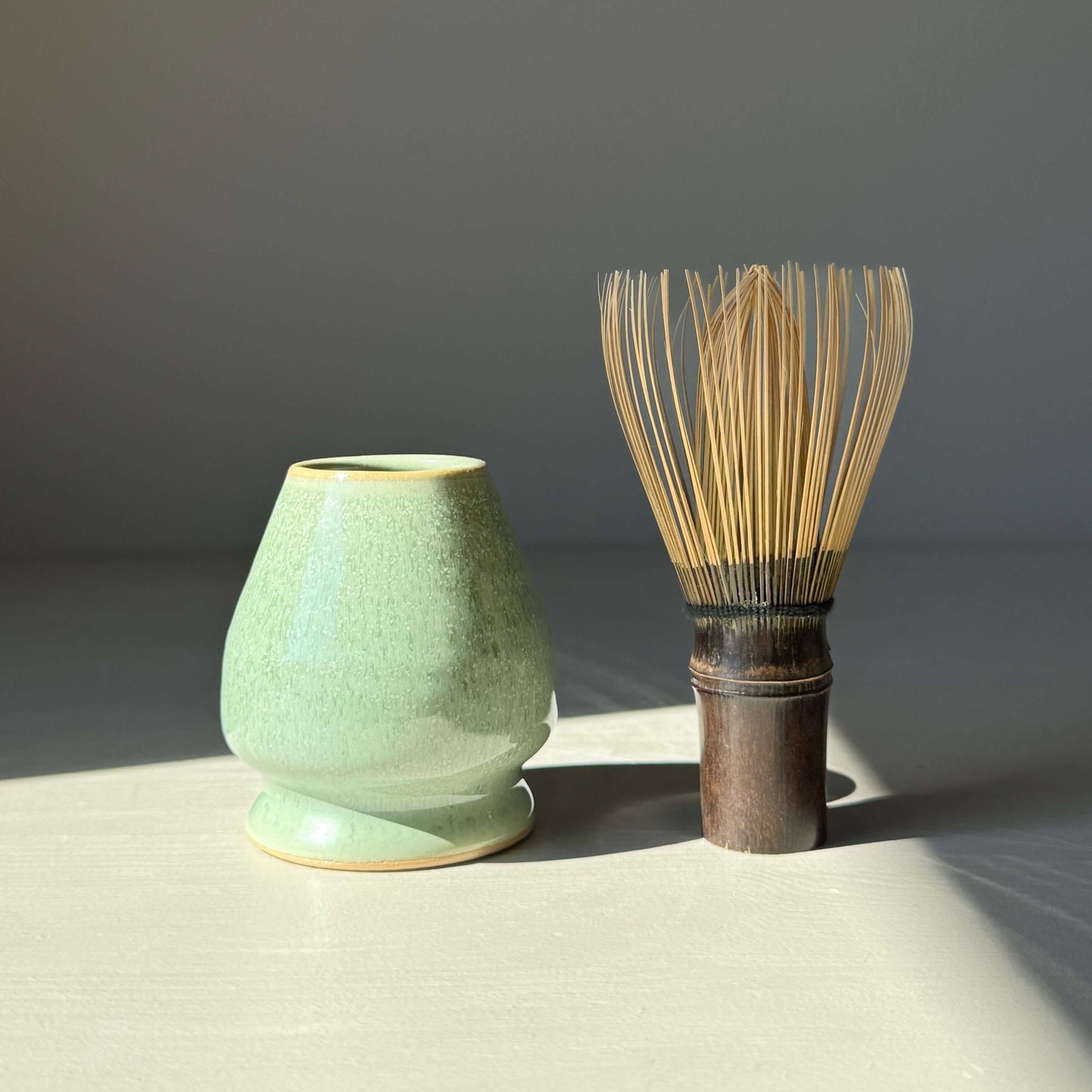 Sage glaze matcha whisk holder with bamboo whisk on a light surface.