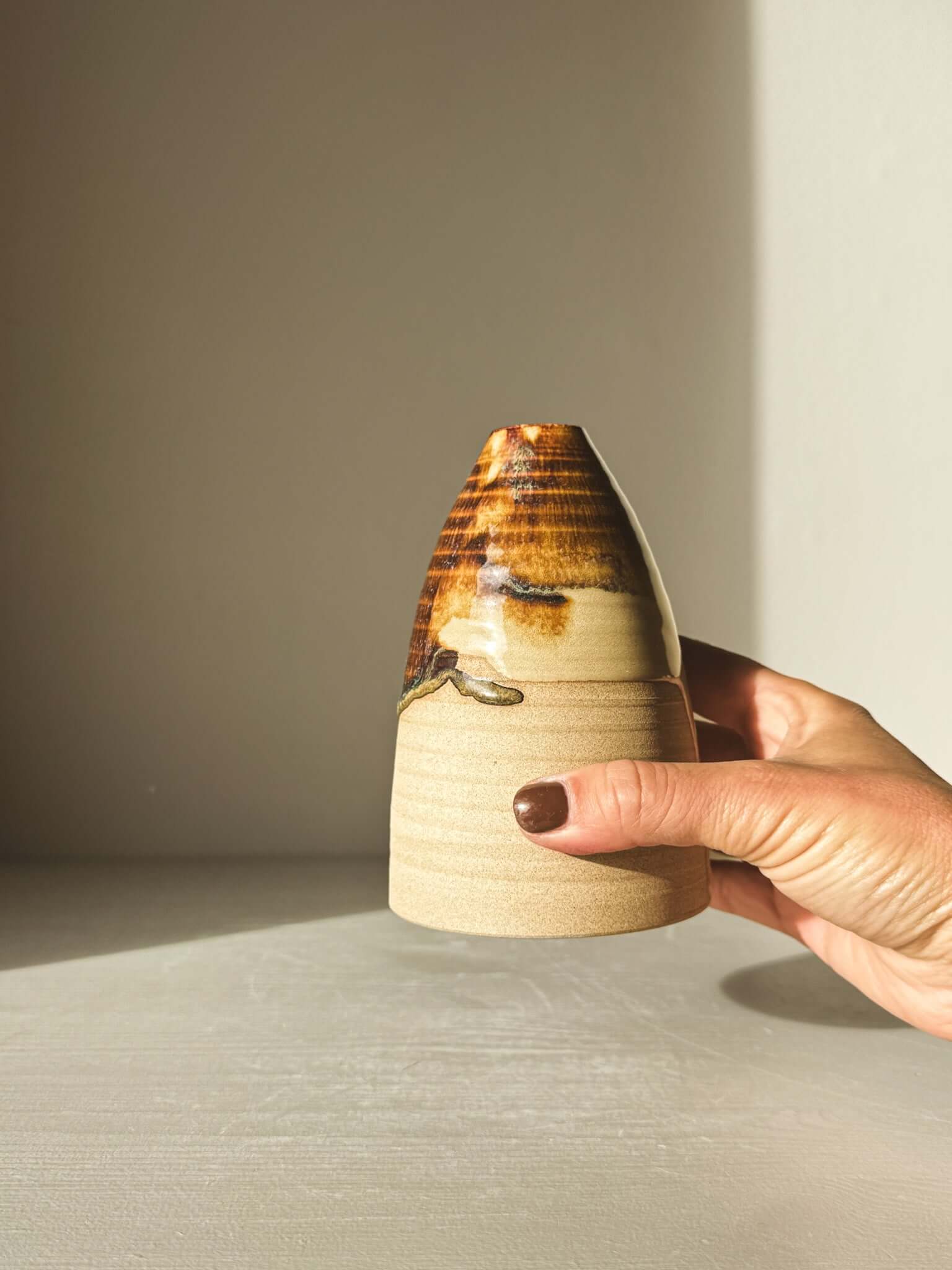 A hand holding the Medium Vase – Inyo #02, showcasing its tenmoku glaze, soft curves, and elegant design in warm lighting.