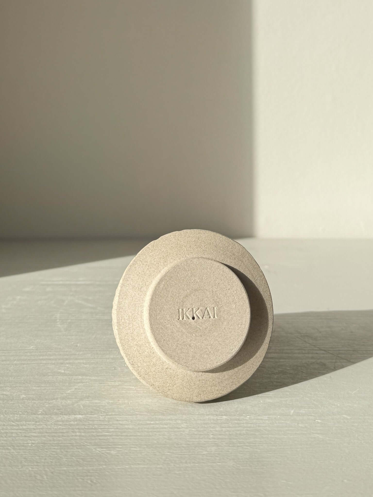 Minimalist ceramic object with engraved letters 'IKKAI', casting shadow on light surface