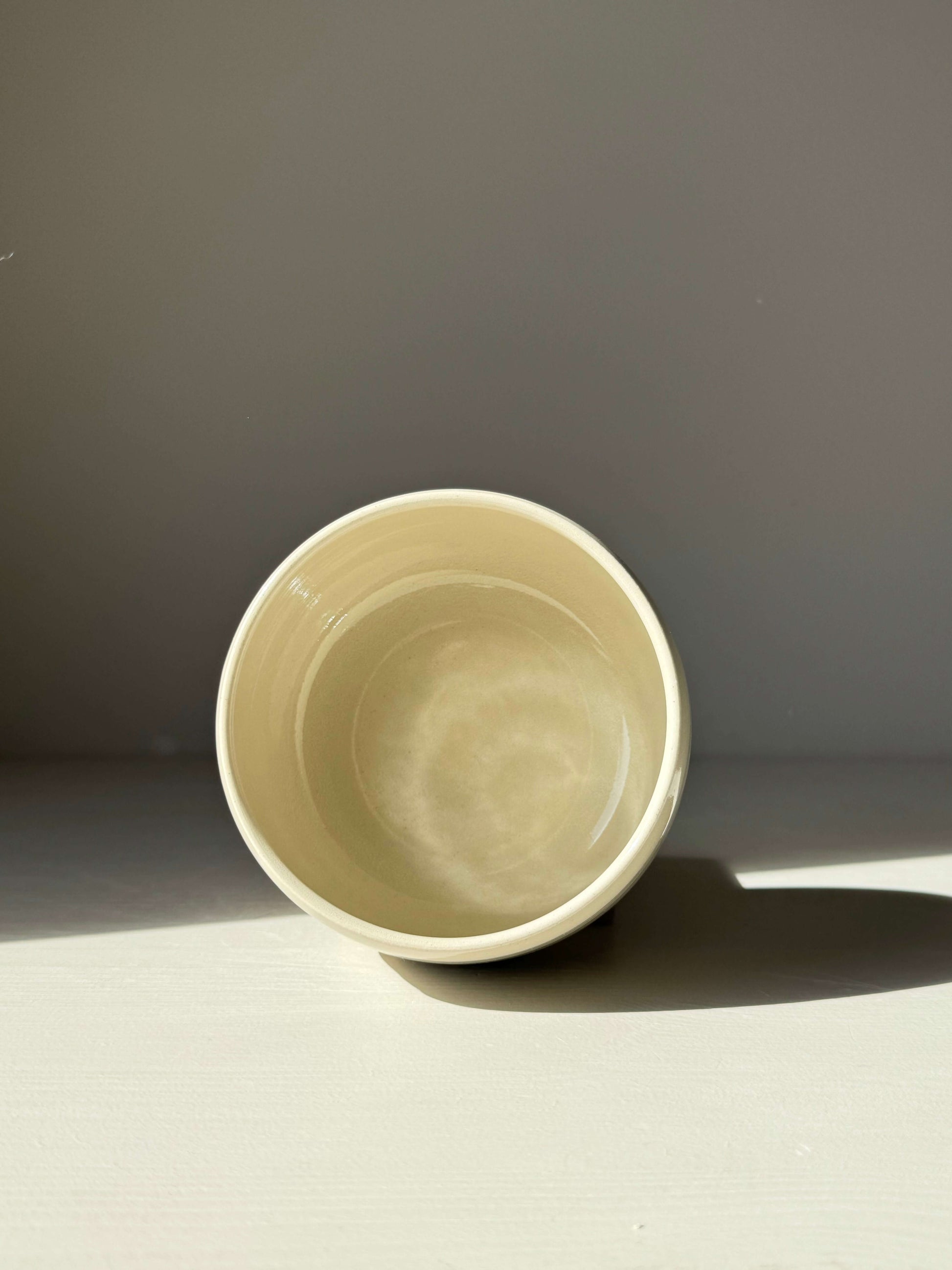 Cream ceramic bowl with sunlight casting a shadow, showcasing a smooth, minimalist design on a subtle gray background.