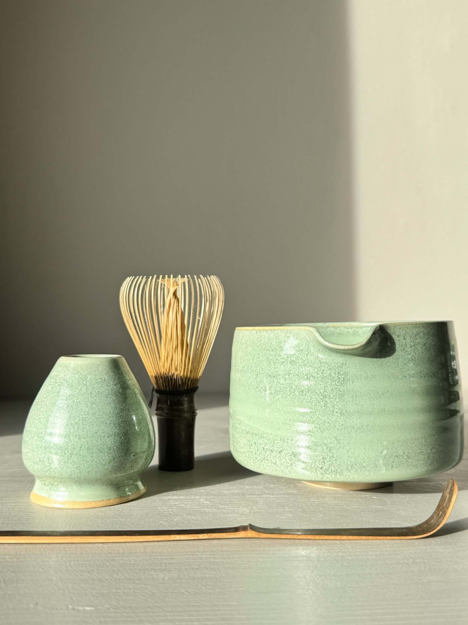 Matcha Bowl Spout – Sage #02 with bamboo whisk and spout resting on a table, showcasing its soft sage-green glaze in sunlight.