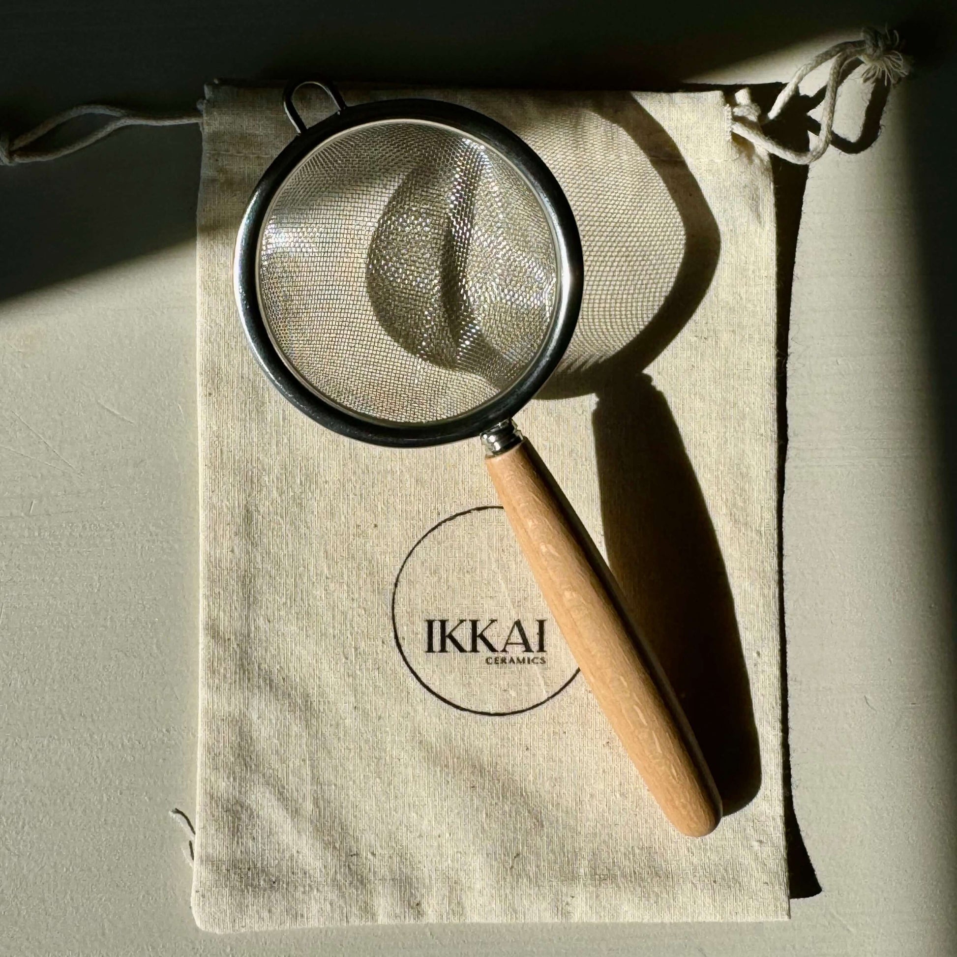 Stainless steel tea strainer with wooden handle on a fabric pouch, perfect for sifting matcha or straining loose leaf tea.