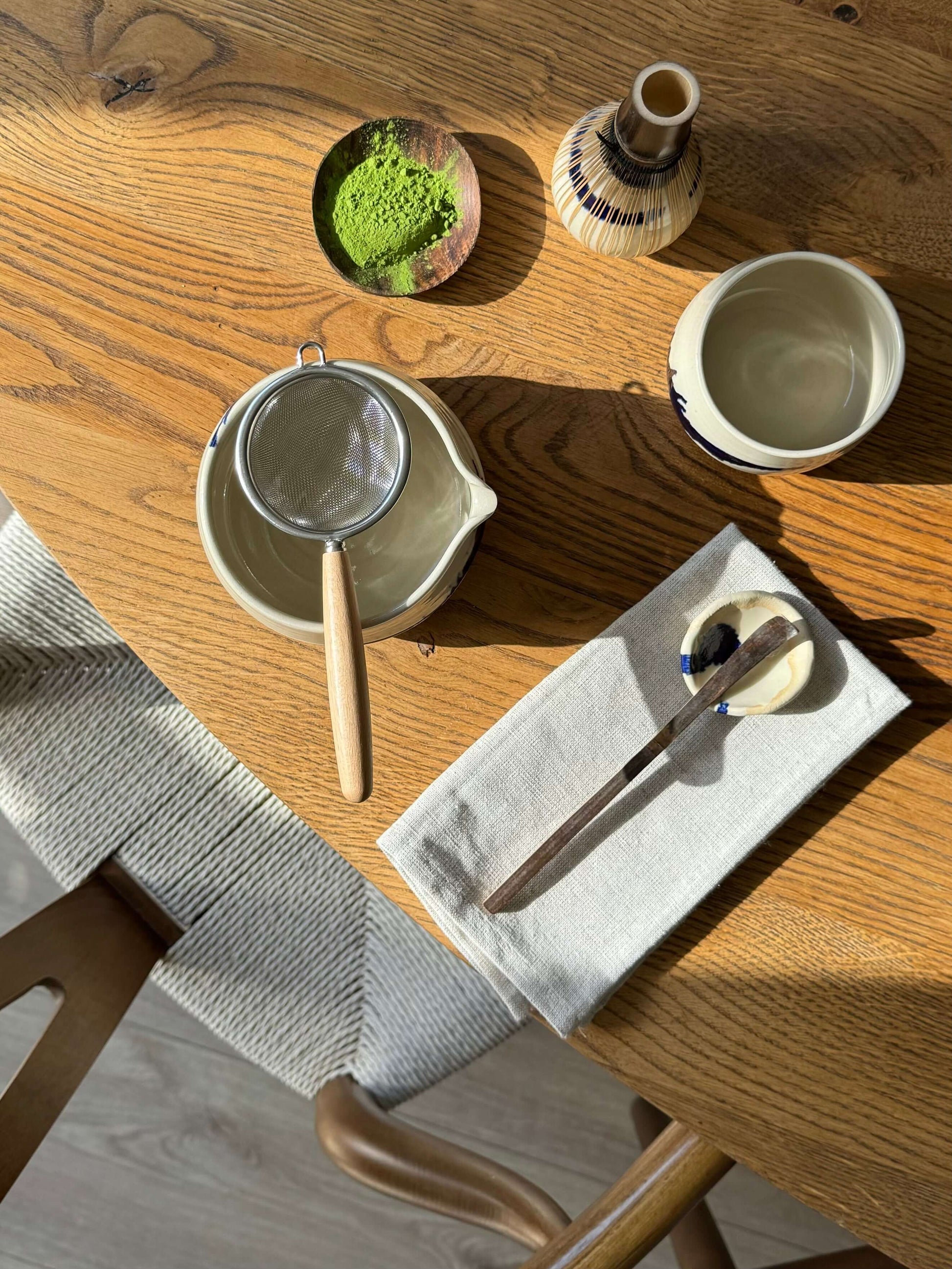 Stainless steel tea strainer with wooden handle on a table, next to matcha powder and ceramic tea set
