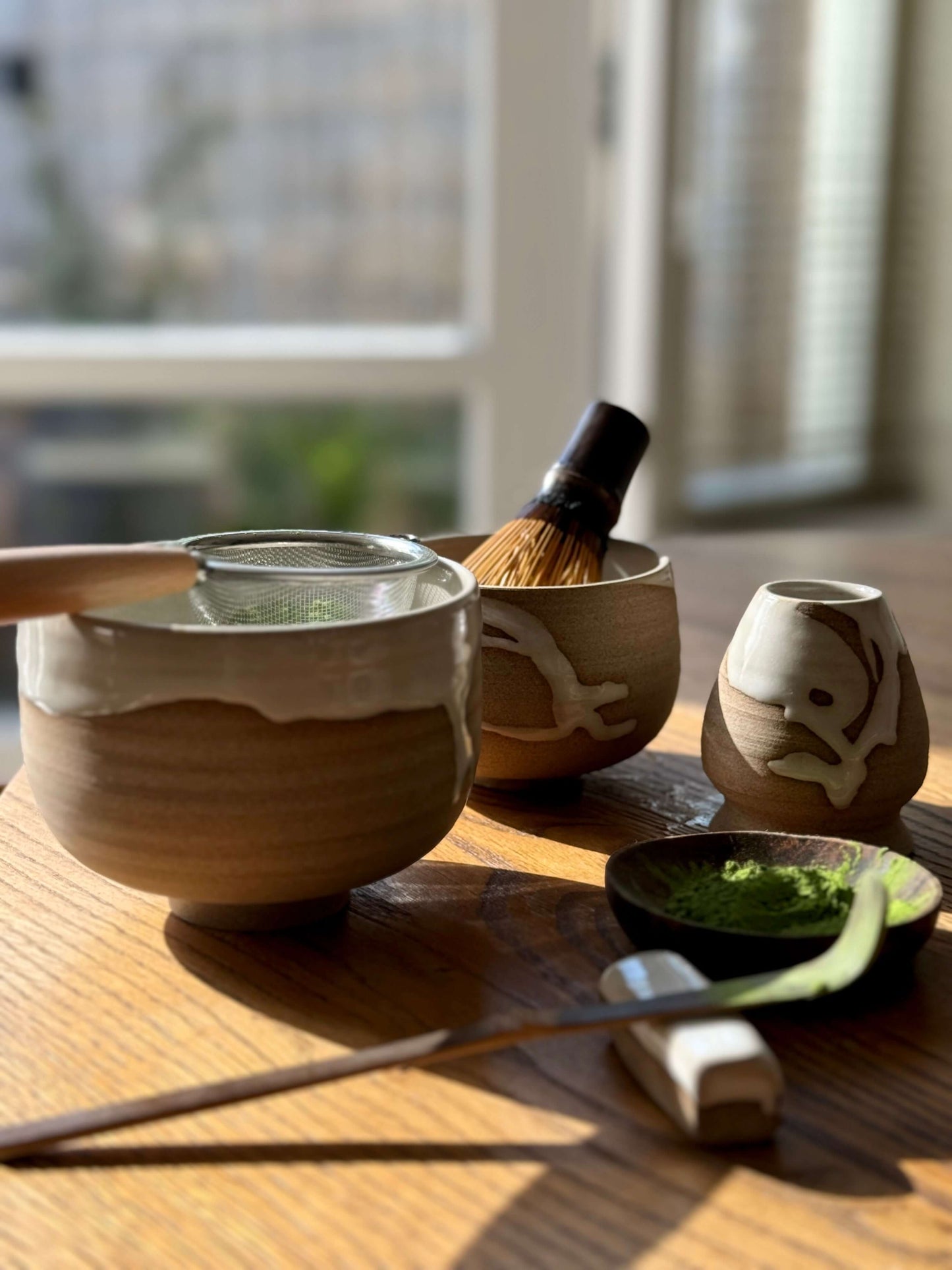 Ceramic bowls with matcha powder, a bamboo whisk, and a strainer on a wooden table, showcasing a serene tea preparation scene.