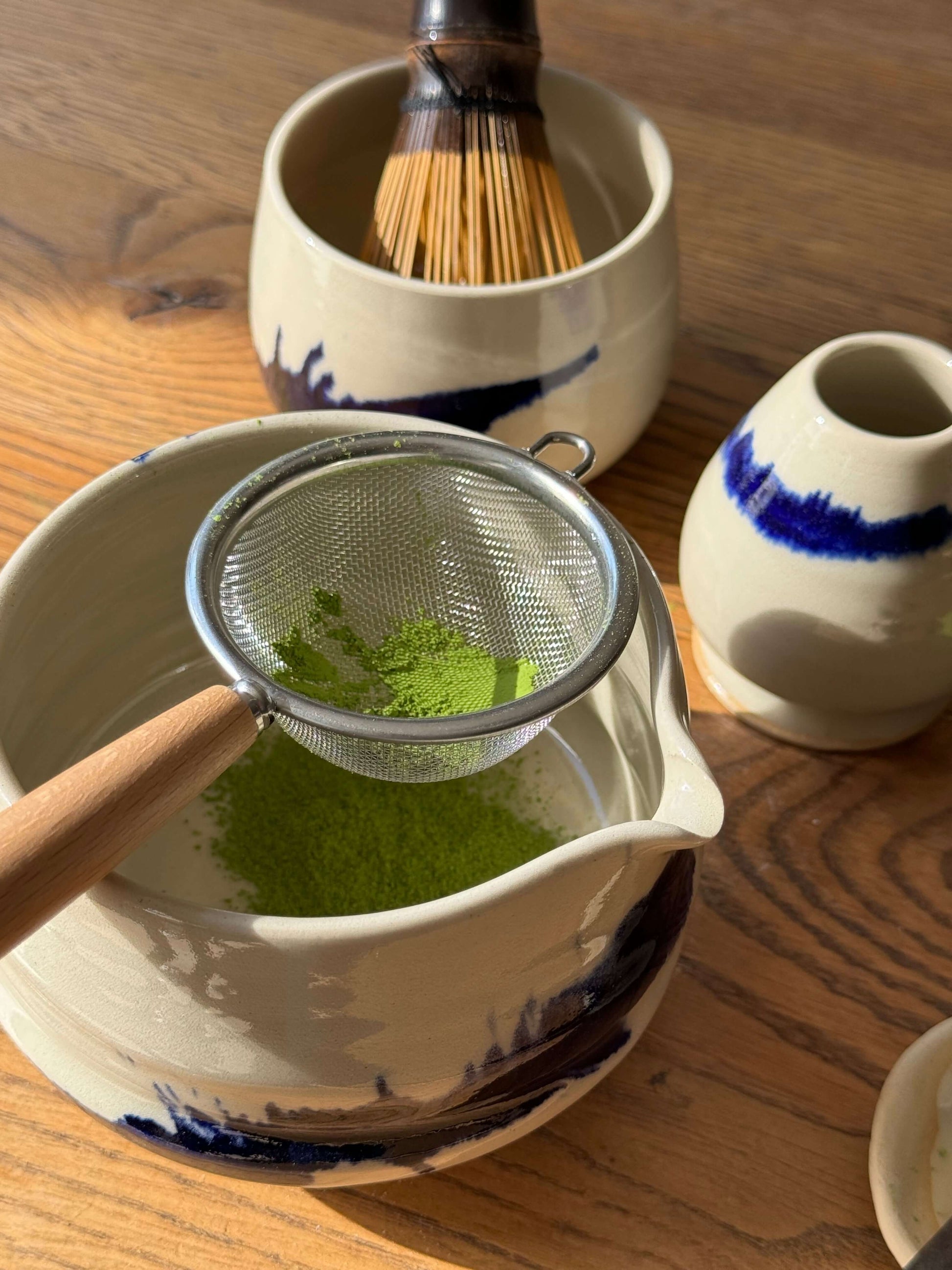 Stainless steel tea strainer with wooden handle sifting matcha powder into ceramic bowl.