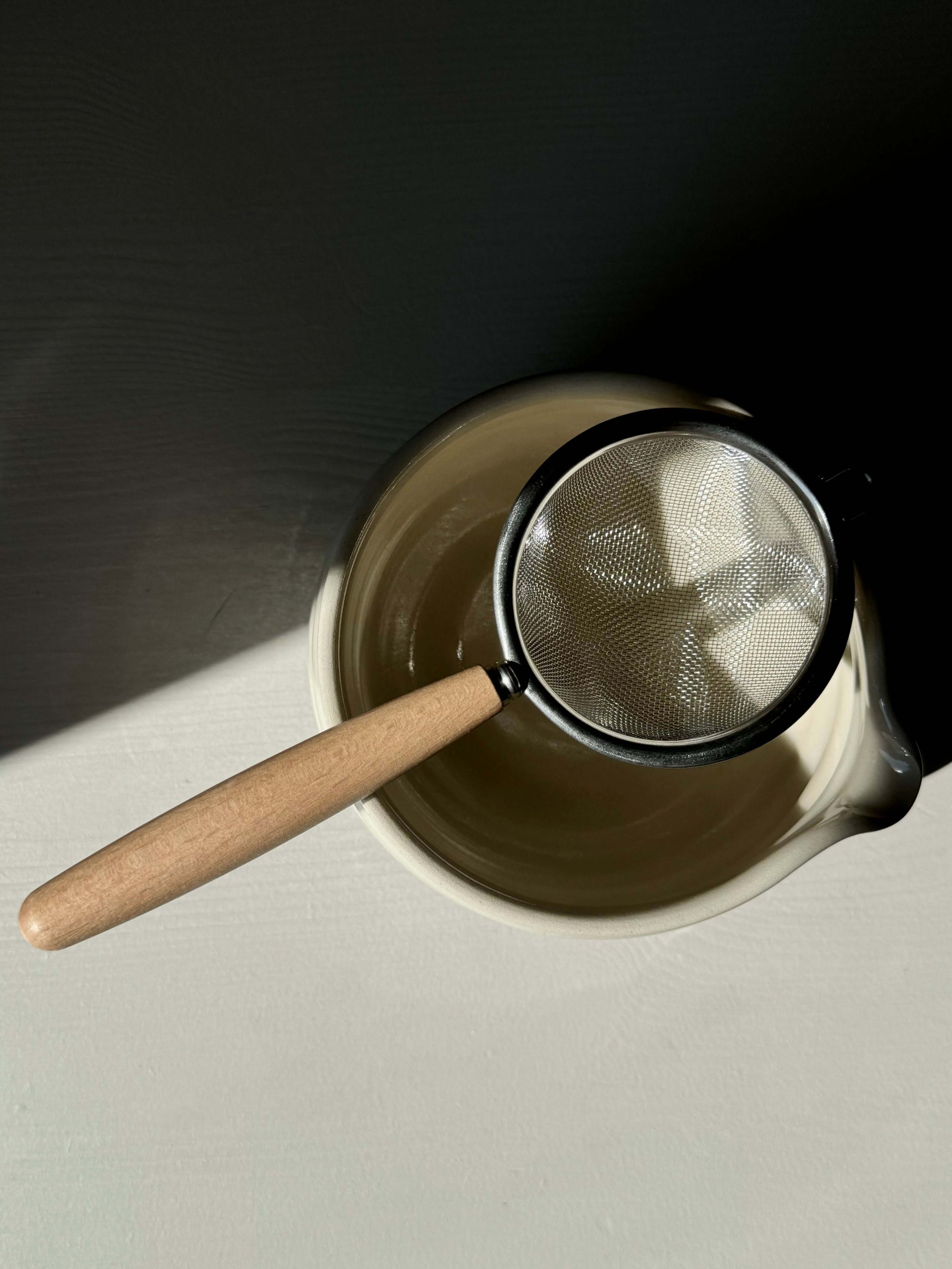 Stainless steel tea strainer with wooden handle resting on a ceramic bowl, perfect for sifting matcha or loose leaf tea.