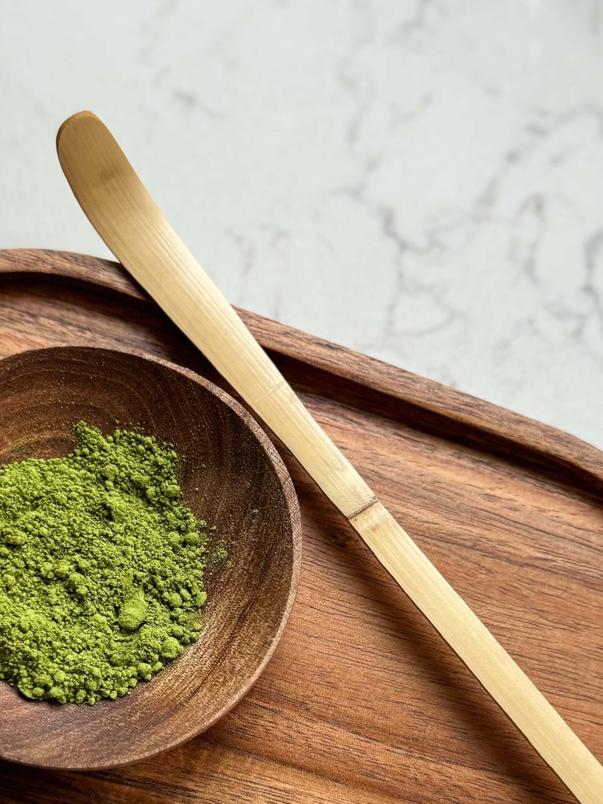 Traditional matcha spoon (Chashaku) made from dark bamboo, placed beside a bowl of matcha powder on a wooden tray.