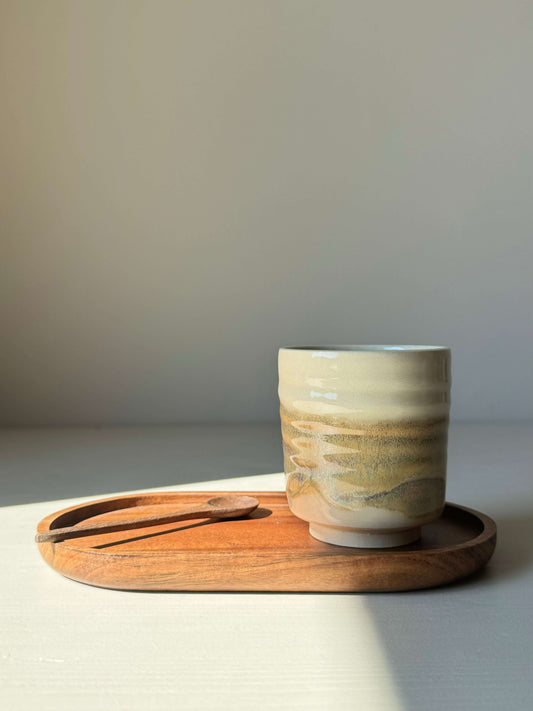 Yunomi cup with clear glaze and tenmoku finish on a wooden tray with a spoon, showcasing handcrafted pottery art.